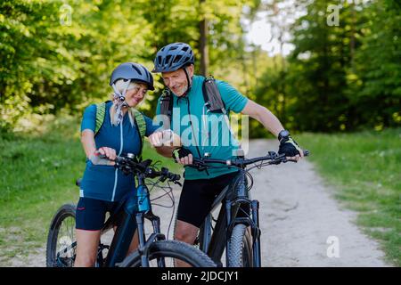 Portrait of active senior couple riding bicycles at summer park, looking at sports smartwatch, checking their performance. Stock Photo