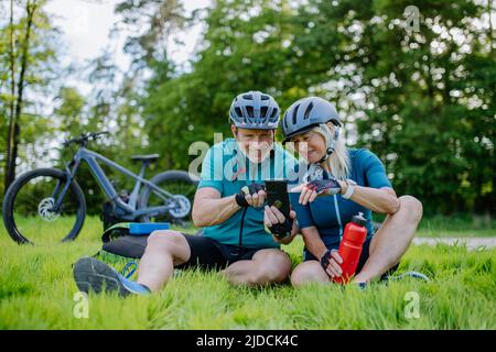 Active senior couple resting after bicycle ride at summer park, sitting on grass and using smartphone. Stock Photo