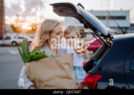 Young mother with little daughter after shopping holding zero waste shopping bags with grocery near car. Stock Photo