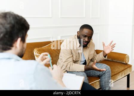 An irritated African American emotionally talks about problems to a psychologist at a session, a confused unhappy young man undergoes therapy with a Stock Photo