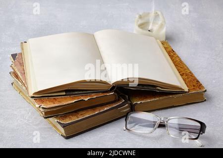 Open vintage notepad book with blank pages and reading glasses lie on a stack of old shabby books against the background of a candle stub on a gray co Stock Photo
