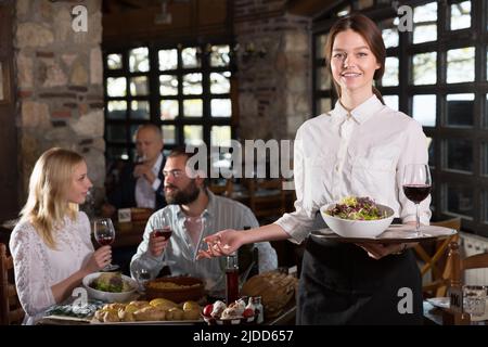 Positive woman waiter demonstrating country restaurant to visitors Stock Photo