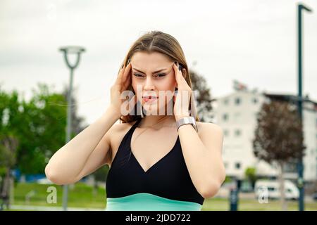 Young caucasian woman wearing black sports bra standing on city park, outdoors holding her head with hands trying to remember something or having head Stock Photo