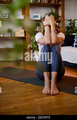 A fit yogi woman sitting on a yoga mat at home with hands-on her eyes and meditating. A yogi woman relaxing at home. Stock Photo