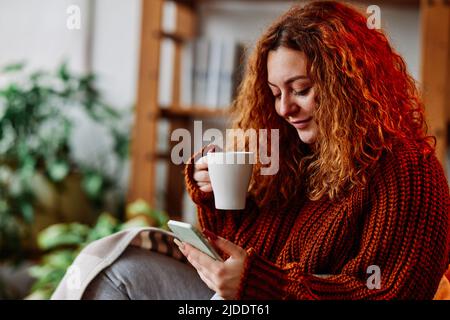 A cute ginger girl with curly hair is sitting in the chair at home in the morning, texting messages on the phone and drinking her coffee. Stock Photo