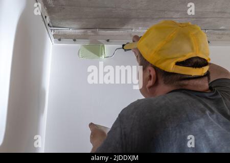 A middle-aged man in dirty clothes and a yellow cap paints a wall with a paint roller. Builder painting a wall in green. Painter painting a wall with Stock Photo