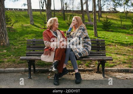 A young ginger woman is sitting with her grandmother and talking on the park bench in the cold weather. Stock Photo