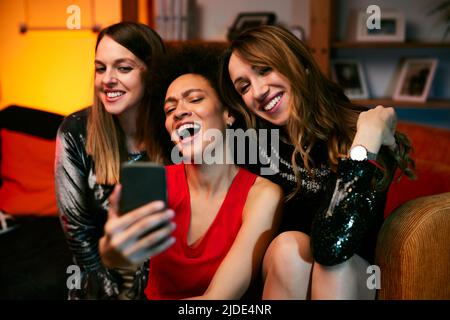Three multicultural female friends sit in the living room in the evening and take selfies at home party. Stock Photo