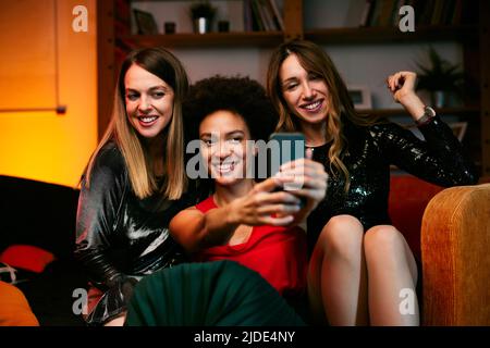 Three multicultural female friends sit in the living room in the evening and take selfies at home party. Stock Photo