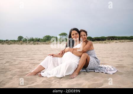 old mother and pregnant adult daughter are seen hugging and smiling on a beach with white dress Stock Photo