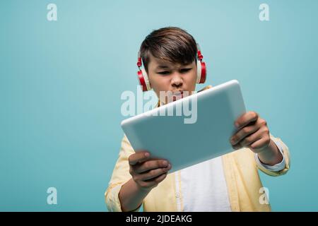 Focused asian schoolkid in headphones using digital tablet isolated on blue Stock Photo
