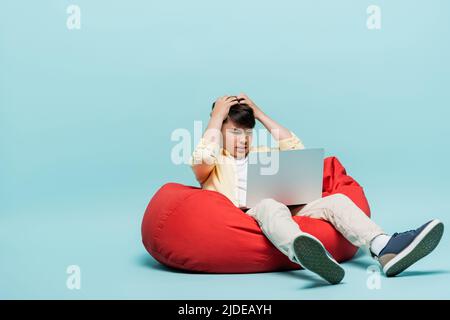 Worried asian schoolkid looking at laptop on beanbag chair on blue background Stock Photo