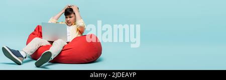 Stressed asian schoolkid looking at laptop on beanbag chair on blue background, banner Stock Photo