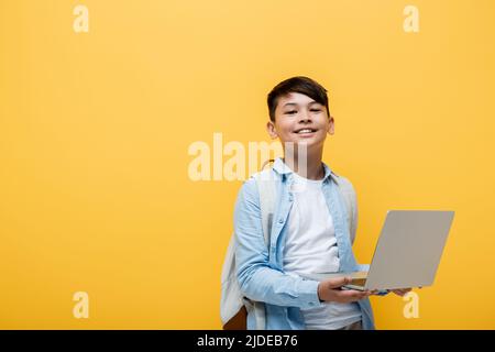 Smiling asian schoolboy holding laptop isolated on yellow Stock Photo