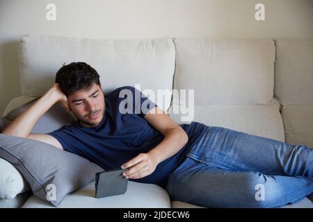 Young man lying down on the sofa reading an ebook Stock Photo