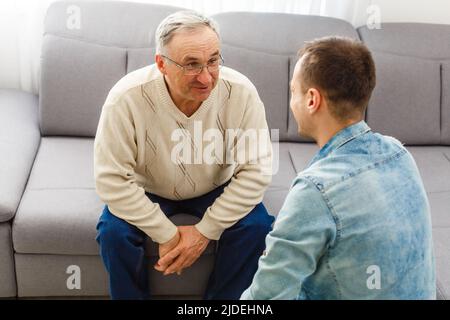 Smiling grown son sit on couch relax with senior dad talk sharing thoughts looking in eyes, happy millennial man rest on sofa speak with elderly Stock Photo