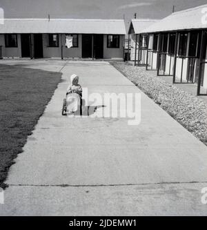 1954, historical, summertime and a young child in a ride-on toy pedal car on a path outside chalets at a holiday camp, which surprisingly is empty of people, England, UK. Stock Photo