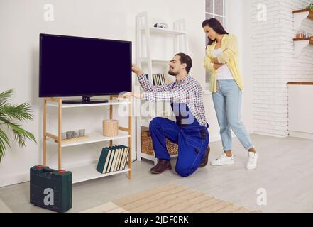 Professional workman in workwear installing large TV monitor in house of female client. Stock Photo