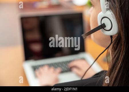 Call center, home office, customer support, help desk concept. Woman with headset working with a laptop, talking on phone, view from behind Stock Photo