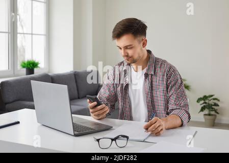 Young businessman using mobile phone while sitting at working desk with laptop computer Stock Photo