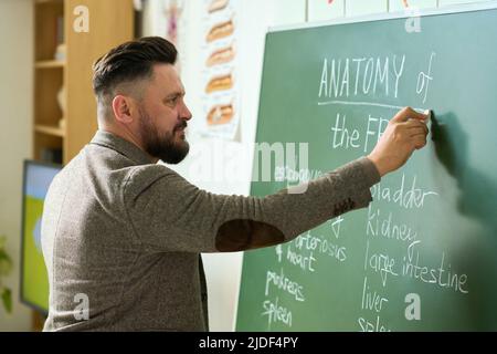 Mature teacher writing down names of frog internal organs on blackboard while explaining students of high school subject of lesson Stock Photo