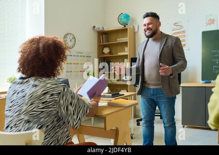 Smiling confident teacher in smart casualwear explaining subject to young female student with open book sitting by desk in front of him Stock Photo