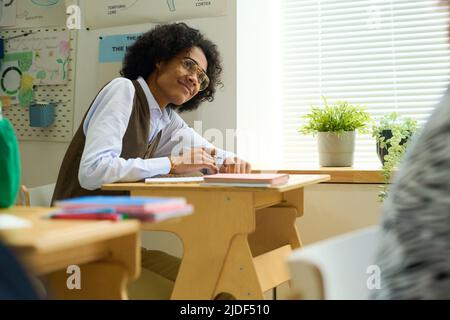 Happy adolescent guy in smart casualwear and eyeglasses sitting by desk in classroom, listening to teacher explanation and making notes Stock Photo