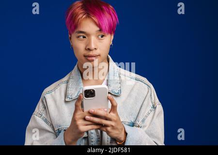 Asian boy in denim jacket looking aside while using mobile phone isolated over blue background Stock Photo