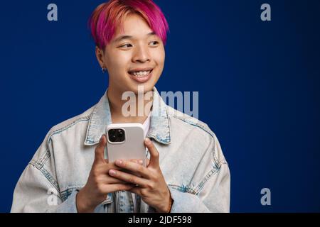 Asian boy in denim jacket smiling while using cellphone isolated over blue background Stock Photo