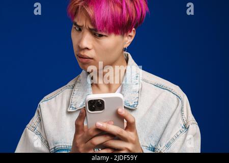 Asian boy in denim jacket frowning while using cellphone isolated over blue background Stock Photo