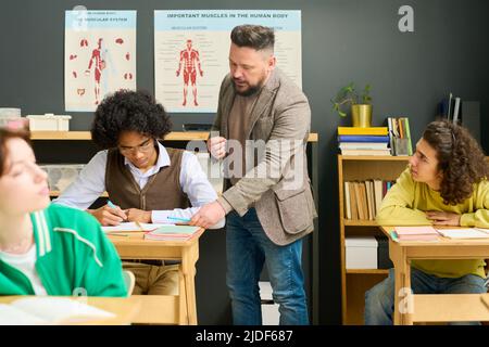 Mature male teacher of anatomy checking notes of highschool student and consulting him among classmates at lesson in classroom Stock Photo