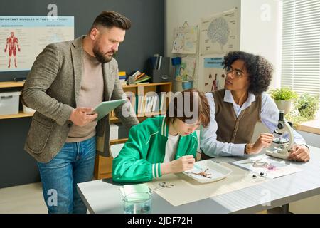 African American highschool student looking at teacher of anatomy while studying dissected parts of frog in microscope at lesson Stock Photo