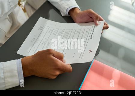 Hands of African American teenage guy holding paper with exam test and looking through assignments while sitting by desk at lesson Stock Photo