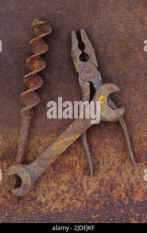 Rusty old double-headed spanner lying next to large drill bit and rusty pliers on rusty metal sheet