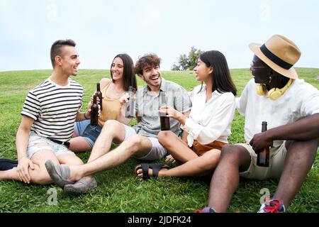 Diverse group of young people celebrating together outdoors sitting on the grass. Friends having fun in summertime.  Stock Photo