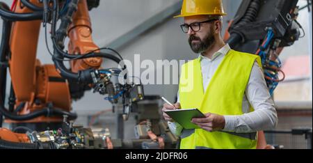 Automation engineer uses tablet for programming robotic arm in factory. Stock Photo
