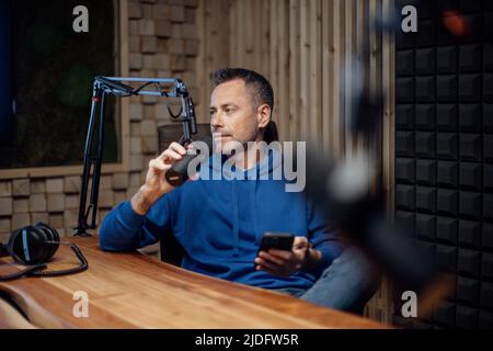 Portrait of mature radio host reading script in cellphone and speaking in microphone while moderating a live show Stock Photo