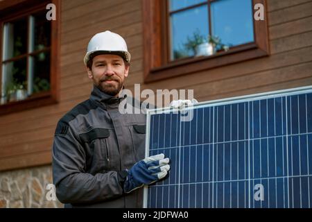 Smiling handyman solar installer carrying solar module while installing solar panel system on house. Stock Photo