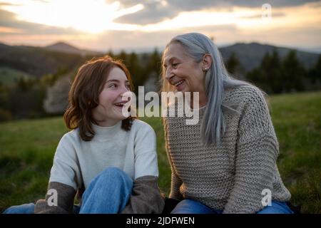 Happy senior grandmother with teenage granddaguhter sitting on grass, talking and having nice time together in nature on spring day. Stock Photo