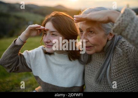 Portrait of happy senior grandmother with teenage granddaguhter sitting on grass and looking into the distance in nature on spring day during sunset. Stock Photo