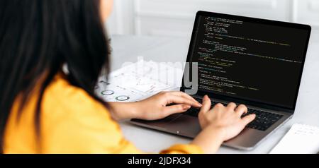 Panoramic over shoulder view female IT-specialist to a laptop with programming code on the screen. Smart professional female programmer working with a software and coding technology, writing codes Stock Photo