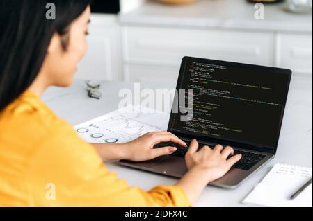 Over shoulder view of asian woman, IT specialist, at a laptop with programming code on the screen. Smart professional female programmer working with a software and coding technology, writing codes Stock Photo