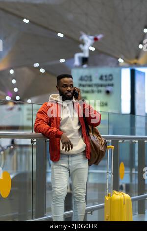 African American guy talking on mobile phone in airport while waiting with suitcase for flight Stock Photo
