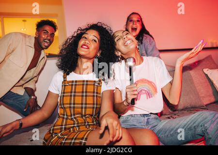 Two young women entertaining their friends on karaoke night. Cheerful girlfriends singing their favourite song at a house party. Group of multicultura Stock Photo