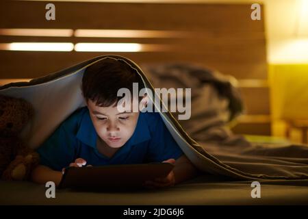 Preteen boy lying under blanket and playing game on tablet computer late at night Stock Photo