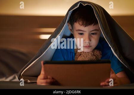 Pensive boy lying under blanket late at night and watching animated cartoon on digital tablet before sleep Stock Photo