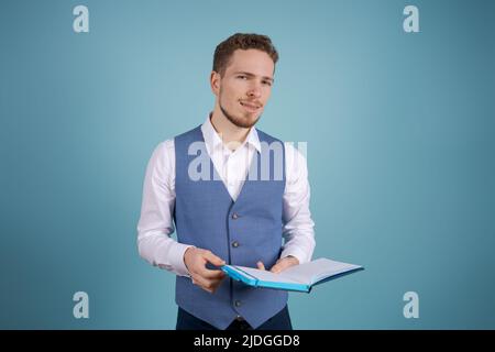 Portrait positive handsome young business assistant with a beard standing on a blue background in business clothes and making notes in a diary Stock Photo