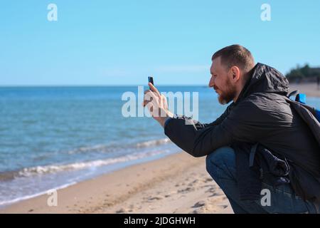 Bearded man in black raincoat on a blue sea landscape background with smartphone in hands talking. Middle aged guy calling friends on the cell phone o Stock Photo