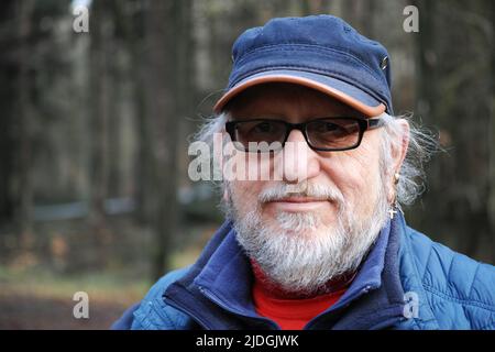 Portrait of a 61-year-old senior man wearing blue clothes and a cap. He has a beard and glasses with tinted lenses Stock Photo