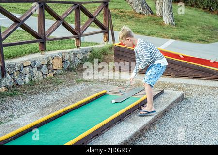 Boy enthusiastically plays mini golf on summer evening on the territory of the hotel Stock Photo